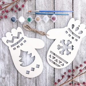 May include: Two unfinished wooden Christmas ornaments shaped like mittens. One mitten has a snowflake design and the other has a Christmas tree design. The ornaments are on a white wooden background with red berries and paint supplies.