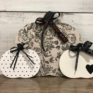 May include: Three wooden pumpkins decorated for Halloween. The largest pumpkin is covered in a black and white floral print fabric and has a black ribbon with the text "BOO!" on it. The other two pumpkins are white with black accents. One has a black heart and the other has a black and white polka dot pattern.