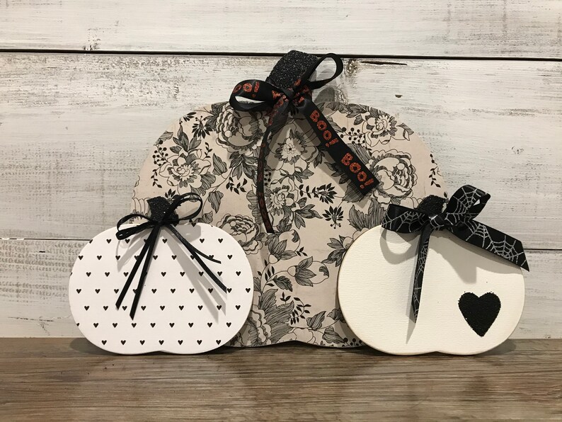 May include: Three wooden pumpkins decorated for Halloween. The largest pumpkin is covered in a black and white floral print fabric and has a black ribbon with the text "BOO!" on it. The other two pumpkins are white with black accents. One has a black heart and the other has a black and white polka dot pattern.
