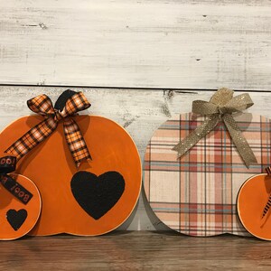 May include: Three wooden pumpkins with different patterns and colors. The largest pumpkin is orange with a black heart and a black and orange plaid ribbon. The middle pumpkin is plaid with a beige, pink, and blue pattern. The smallest pumpkin is orange with black stripes and a black heart.