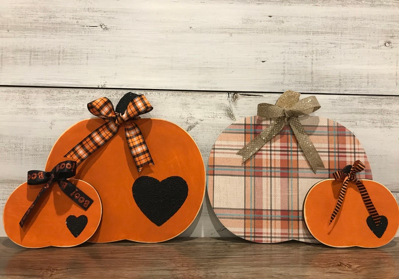 May include: Three wooden pumpkins with different patterns and colors. The largest pumpkin is orange with a black heart and a black and orange plaid ribbon. The middle pumpkin is plaid with a beige, pink, and blue pattern. The smallest pumpkin is orange with black stripes and a black heart.