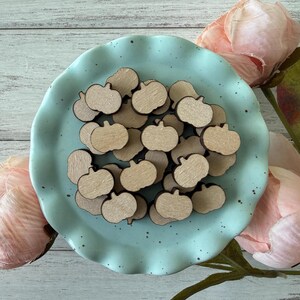May include: A collection of small, wooden pumpkin cutouts arranged on a light blue, ruffled-edge plate. The pumpkins are a natural wood color and are scattered across the plate. Pink flowers are in the background.