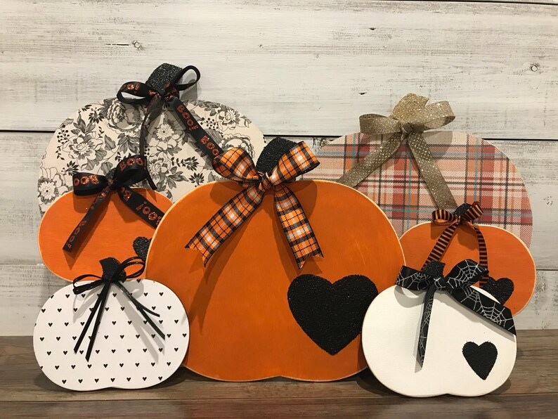 May include: Five wooden pumpkins decorated for Halloween. The pumpkins are various sizes and feature different patterns, including plaid, floral, and polka dots. Each pumpkin has a black heart and a black ribbon.