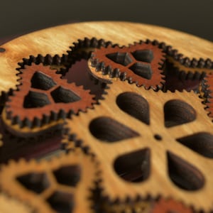 May include: A wooden circular ornament with intricate cut-out designs. The ornament features a central flower-shaped design surrounded by smaller, gear-like shapes. The wood is a light brown color with darker brown accents.