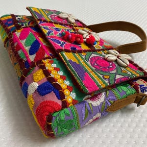 Puede incluir: Un bolso colorido estilo patchwork con asa marrón. El bolso presenta bordados vibrantes en rosa, verde, morado y amarillo, adornado con conchas marinas y pompones rojos. El diseño del bolso es una mezcla de patrones florales y geométricos.