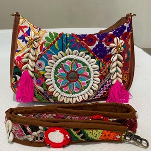 Puede incluir: Bolso de hombro patchwork colorido con ribete y correa de cuero marrón. El bolso presenta un diseño circular central con un espejo, rodeado de conchas blancas y cuentas. Borlas rosas y detalles de conchas añaden detalles.