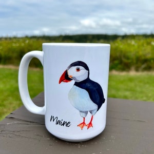 May include: White ceramic mug featuring a watercolor illustration of a puffin bird with a red beak, black and white plumage, and orange feet. The word "Maine" is written in black script below the bird. The mug has a large handle.
