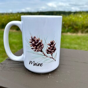 May include: White ceramic mug with a brown pine cone design and the word "Maine" printed on the front.