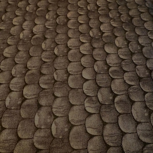 Puede incluir: Primer plano de una tela con un patrón repetitivo de formas redondeadas superpuestas, que recuerdan a escamas. Las escamas son en tonos marrones y grises, creando una apariencia texturizada y orgánica. La tela tiene un efecto sutil y moteado.