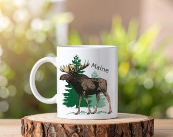 May include: White ceramic mug featuring a brown moose illustration with green trees and the word "Maine" in a script font. The mug is on a wooden slice, with a blurred green background.