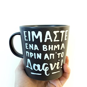 May include: A black ceramic mug with white lettering that reads "Είμαστε ένα βήμα πριν από το Δαχτυλι!"