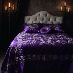 Op de afbeelding: Een luxe bed met een gecapitonneerd hoofdbord en een dieppaarse fluwelen dekbedovertrekset. Het beddengoed heeft een ingewikkeld bloemmotief in een lichtere tint. Kandelaars flankeren het bed en werpen een warme gloed.