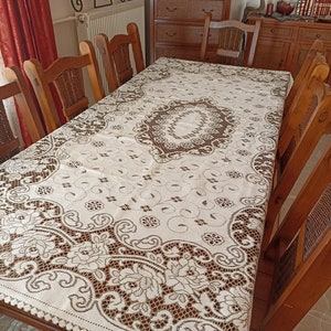 May include: A white tablecloth with a brown lace pattern. The tablecloth is laid on a wooden table with chairs around it.