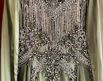 May include: An olive green long-sleeved dress featuring a high neckline and intricate beadwork across the chest. The bodice is adorned with silver and pink beads, sequins, and dangling fringe elements that create a detailed, symmetrical pattern.