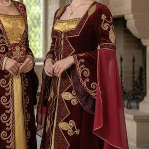 May include: Two women wearing medieval-style gowns. The gowns are primarily burgundy with gold embroidery and accents. One gown has a gold bodice and skirt. The other gown has a black lace skirt and burgundy sleeves with red chiffon accents.