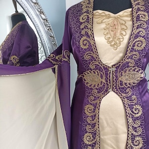 May include: A formal purple dress with gold embroidery and a cream-colored underdress. The dress features intricate gold detailing along the edges and bodice, with long sleeves and a flowing design. The dress is displayed on a mannequin.
