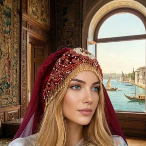 Può includere: Un copricapo di velluto rosso con intricate perline e frange dorate. Il copricapo è adornato con perle e altri elementi decorativi. La donna indossa un velo rosso e un top a fantasia floreale. Lo sfondo presenta una vista panoramica di acqua ed edifici.