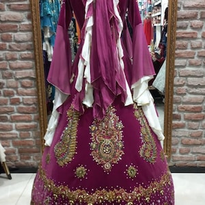 May include: A magenta-colored, ornate dress with long sleeves and a full skirt. The dress features gold embroidery and embellishments, with white accents on the bodice and sleeves. The dress is displayed on a mannequin in front of a mirror.