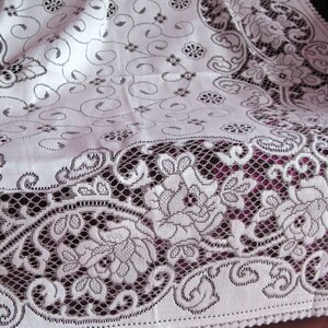May include: White lace tablecloth with a delicate floral pattern. The lace is intricate and has a scalloped edge. The tablecloth is draped over a purple surface.