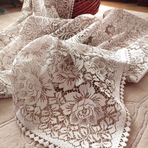 May include: White lace table runner with a delicate floral pattern. The runner has a scalloped edge and is made of a fine, lightweight fabric.