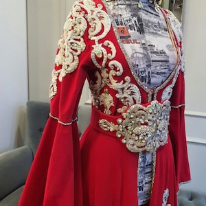 May include: A vibrant red formal gown with elaborate gold and silver embellishments. The dress features a detailed bodice, a wide belt, and flared sleeves. The ensemble includes a matching headpiece, creating a regal and ornate appearance.