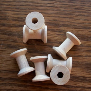 May include: A collection of unfinished wooden spools, ideal for crafting and sewing projects. The spools are made of light-colored wood and have a classic design with a central hole. They are arranged on a wooden surface.
