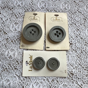 May include: Three sets of gray La Mode buttons on their original cards. The buttons are various sizes and have four or two holes. The cards are off-white and have the La Mode logo.