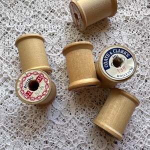May include: A collection of vintage wooden spools, some with labels. The spools are a light tan color, with labels in red, white, and blue. The labels include the text "BOILFAST" and "COATS & CLARK'S". The spools are arranged on a white lace surface.