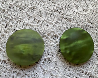 May include: Two round, green earrings with a marbled effect. The earrings are set against a white, lace background. The earrings are a solid color with a slightly translucent appearance.