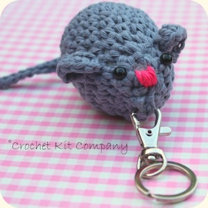 May include: A crocheted gray cat keychain with a pink nose and black eyes. The keychain has a silver clasp and ring. The text "Crochet Kit Company" is in the image.