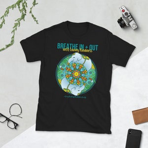 May include: Black t-shirt with a circular graphic featuring two light blue elephants and a colorful mandala design. The text "Breathe In + Out with Loving Kindness" is printed above the graphic. A camera and other items are in the background.
