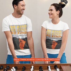 May include: Two people wearing white t-shirts with the text "Be Kind. Be Kind. Be Kind. You're Always Right." and a photo of a person in an orange robe. The text is in yellow and orange. The people are standing in front of a foosball table.