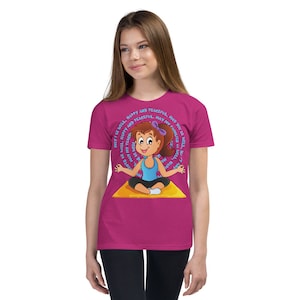 May include: A pink youth t-shirt with a cartoon girl meditating on a yellow mat. The text around the girl says "May I be well, happy and peaceful. May you be well, happy and peaceful. May my teacher be well, happy and peaceful. May my family be well, happy and peaceful."