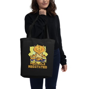May include: A black tote bag with a cartoon cat meditating in a yellow lotus flower. The text "Heavily Meditated" is printed in gold below the cat.