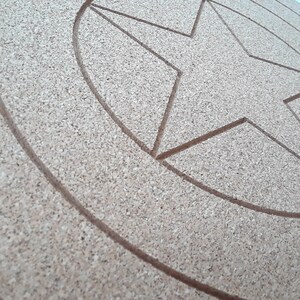 May include: A round, cork-board shield with a five-pointed star design carved into the surface. The shield has concentric circles around the star, and the cork material has a natural, light brown color.