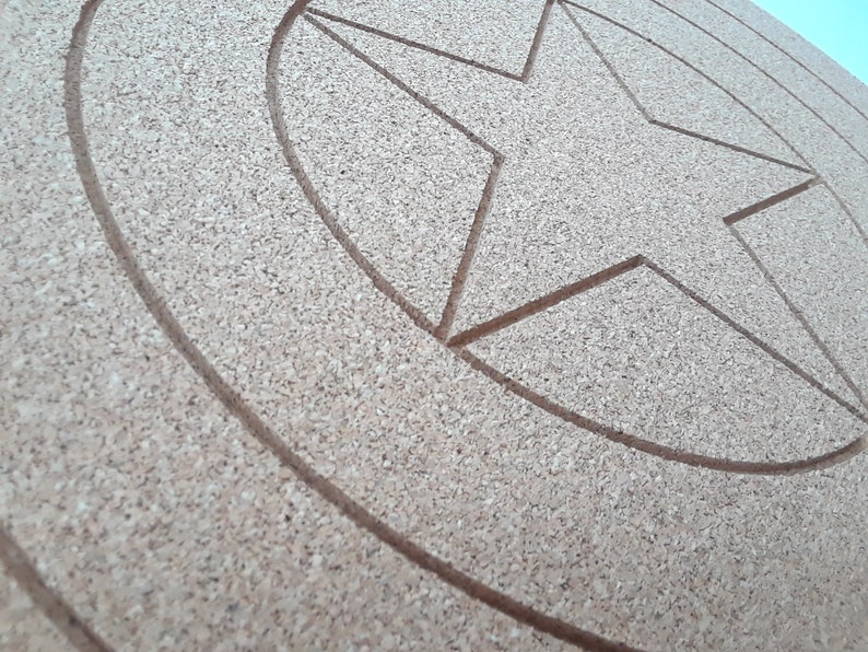 May include: A round, cork-board shield with a five-pointed star design carved into the surface. The shield has concentric circles around the star, and the cork material has a natural, light brown color.