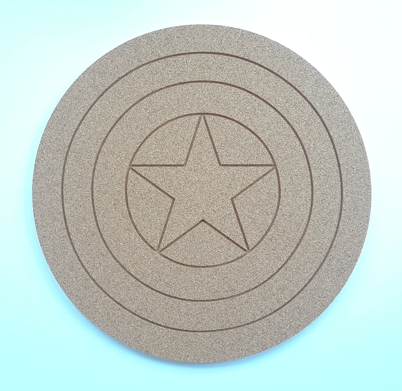 May include: A round, tan-colored coaster featuring a star design in the center, surrounded by concentric circles. The coaster is made of a textured material, resembling cork or wood, and is likely used for placing drinks.