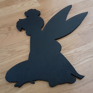 May include: Black wooden cutout of a fairy with wings spread out. The fairy is in a sitting position with her legs crossed.