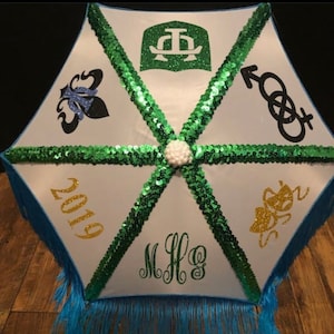 May include: A white parasol with green sequin accents and a blue fringe. The parasol features various glitter designs, including a fleur-de-lis, a crest, and the year 2019. The initials "MHG" are also displayed.