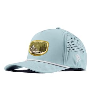 May include: A light blue baseball cap featuring a white cord across the brim and a rectangular patch on the front. The patch displays a mountain and forest graphic with the text PEAK PERF. The side panels have laser-cut ventilation holes.