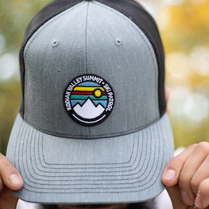 May include: A gray and black trucker hat with a patch that says "Kodiak Valley Summit - Ski Patrol" with a mountain and sun graphic.