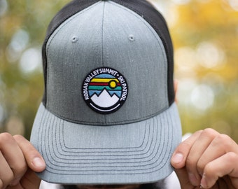 May include: A gray and black trucker hat with a patch that says "Kodiak Valley Summit - Ski Patrol" with a mountain and sun graphic.