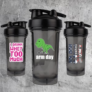 May include: Three black shaker bottles with different designs. The first bottle has the text "I drink whey too much" in pink letters. The second bottle has a green dinosaur lifting a weight with the text "arm day". The third bottle has the text "Arm High Rep Count Low & Slow" with an American flag design.