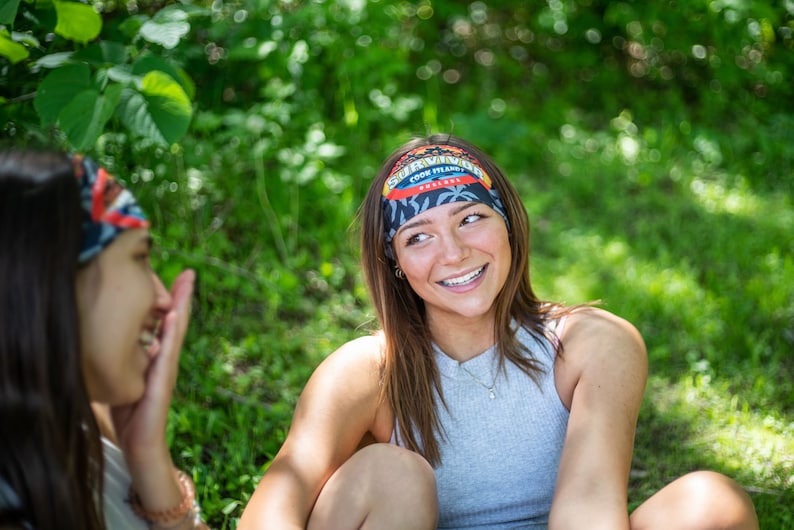 K&ouml;nnte beinhalten: Zwei Personen tragen Stirnb&auml;nder mit dem Text "Survivor Cook Islands" und einem tropischen Design. Die Stirnb&auml;nder sind schwarz, rot und blau. Eine Person tr&auml;gt ein graues Tanktop und l&auml;chelt. Der Hintergrund ist gr&uuml;n.