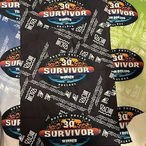 May include: Black fabric with a repeating pattern of white tribal designs. The fabric features a large, colorful logo for the reality TV show "Survivor" with the text "Outwit Outplay Outlast" and "Winner" in a white circle. The logo also includes a sunset and palm trees.
