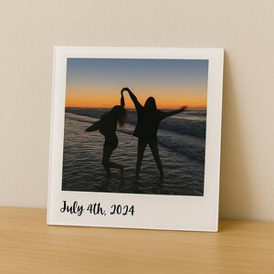 May include: A framed photograph featuring two silhouetted figures dancing in the ocean at sunset. The sky transitions from orange to blue. The text "July 4th, 2024" is printed at the bottom of the photo.