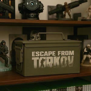 May include: An olive green metal box with a handle and the words "ESCAPE FROM TARKOV" printed in white. The box is rectangular and has a hinged lid. The box is sitting on a wooden shelf with other figurines.
