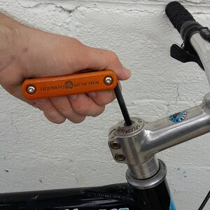 Peut inclure: Une personne utilise un multi-outil orange et noir pour serrer une potence de v&eacute;lo en argent. L'outil porte l'inscription "HIGHROAD MULTI TOOL".