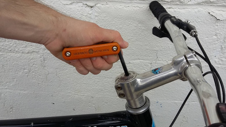 Peut inclure: Une personne utilise un multi-outil orange et noir pour serrer une potence de v&eacute;lo en argent. L'outil porte l'inscription "HIGHROAD MULTI TOOL".