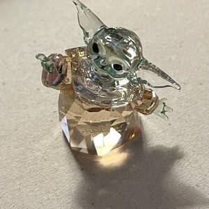 Puede incluir: Una figura de cristal transparente de un personaje con orejas y ojos grandes, sobre una base facetada. La figura tiene los brazos extendidos y est&aacute; hecha de m&uacute;ltiples piezas de cristal. La base es de color &aacute;mbar claro.